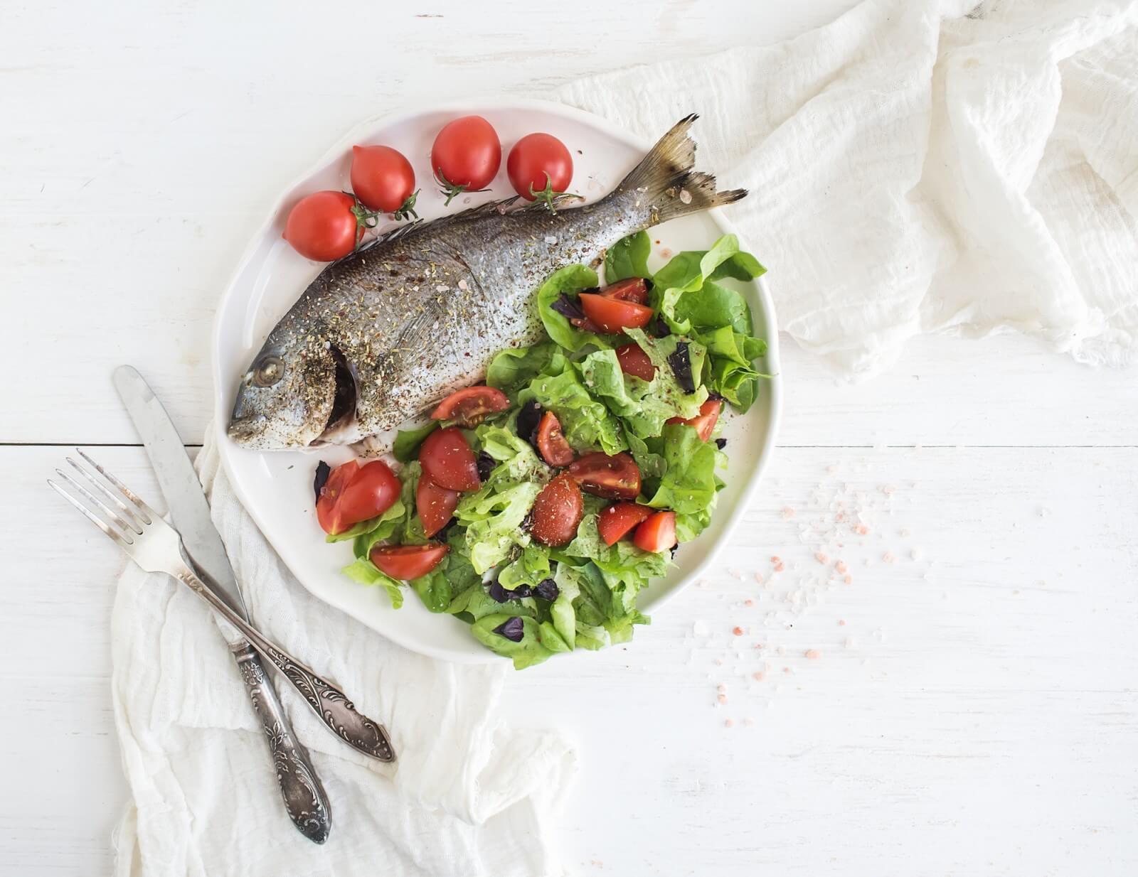 Cooked sea bream fish with fresh vegetable salad on ceramic plate over white rustic wooden backdrop, top view, copy space